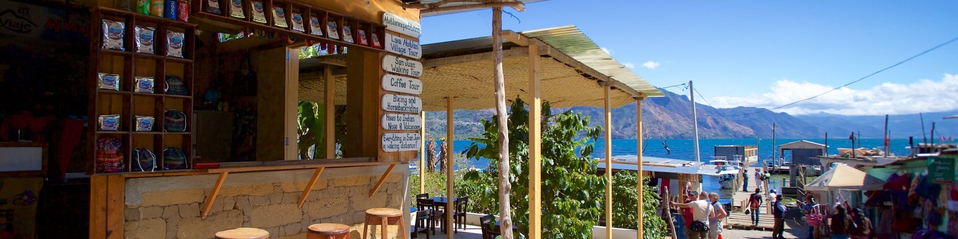 Um café rústico ao ar livre, com banquetas de madeira e balcão de pedra, com vista para um corpo de água e montanhas.