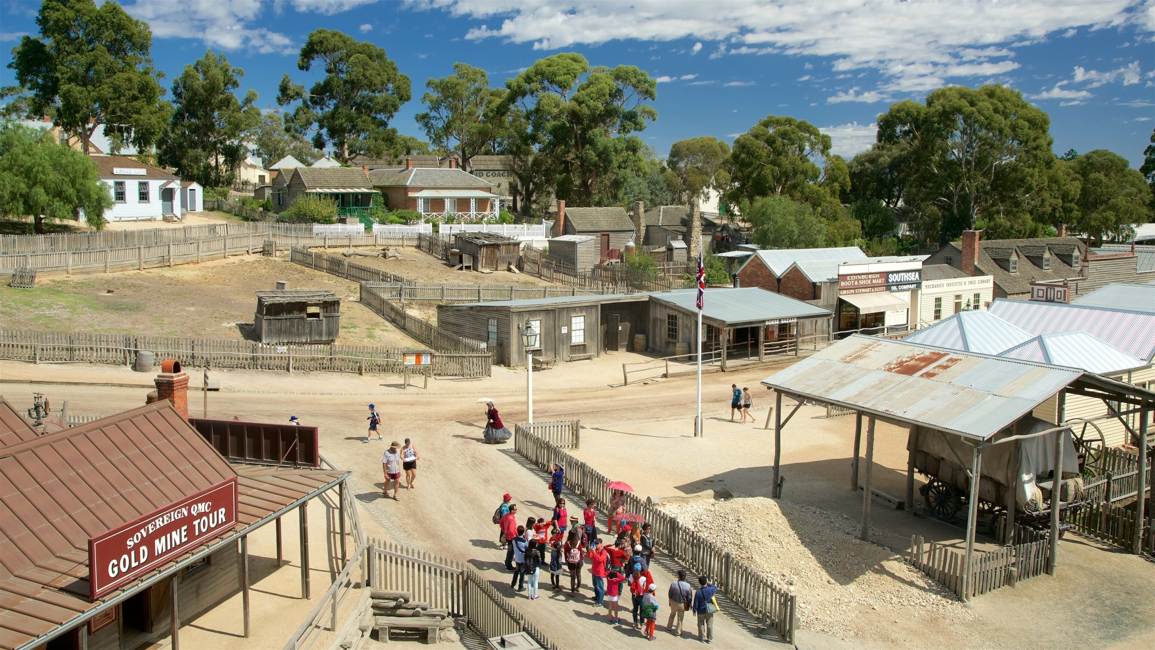 A historical town with wooden buildings, a dirt road, and a fenced area with people.