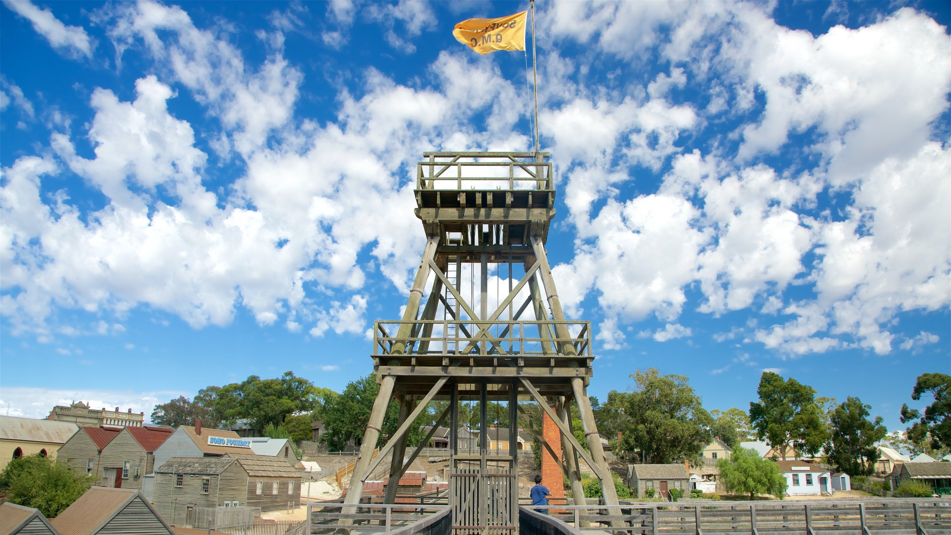 A historic mining tower with a flag, surrounded by old buildings and trees.