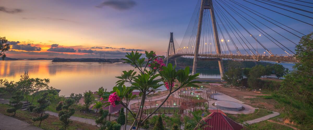 A suspension bridge at sunset with a calm river, greenery, and a small pavilion.