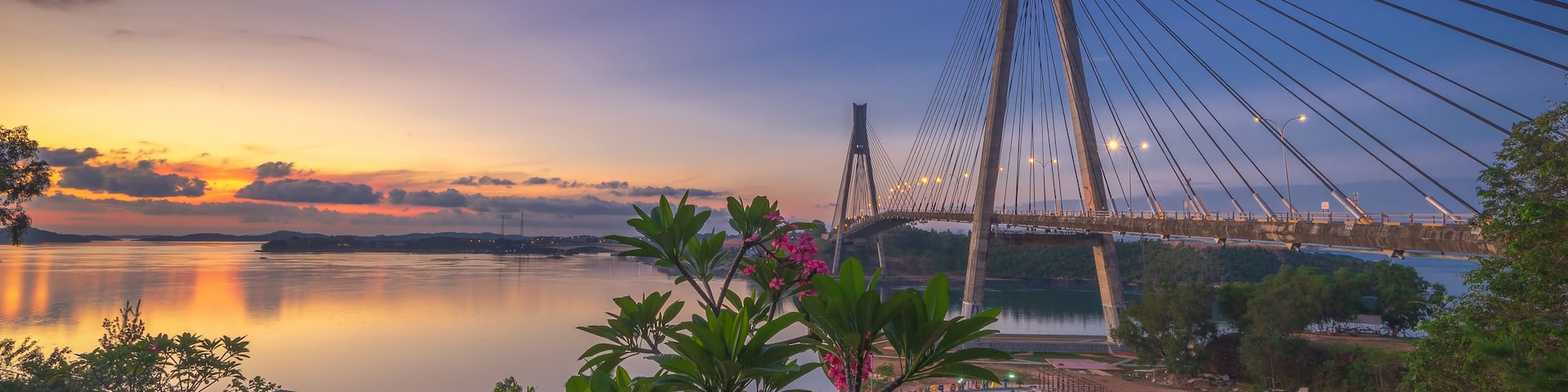 A suspension bridge at sunset with a calm river, greenery, and a small pavilion.
