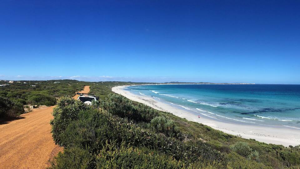 A coastal road leading to a beach with clear blue waters and green vegetation.