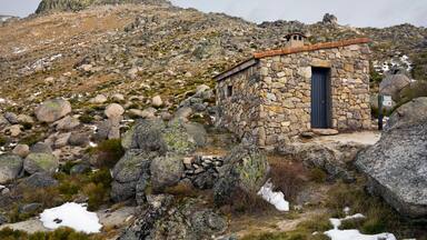 A stone building with a blue door, surrounded by rocks and sparse vegetation on a mountainous terrain.