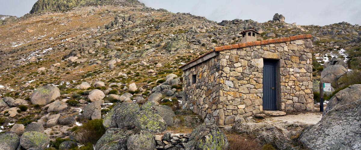 A stone building with a blue door, surrounded by rocks and sparse vegetation on a mountainous terrain.