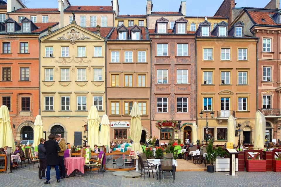 A European street with colorful buildings, outdoor cafes, and people sitting at tables.