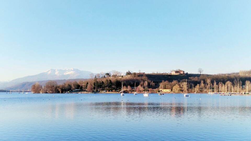 A calm lake with sailboats, a forested hill, and snow-capped mountains in the background.