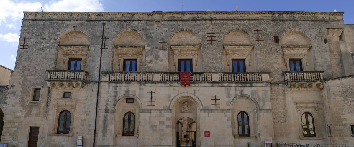 A historic building with arched windows and balconies, a red banner, and a paved courtyard.