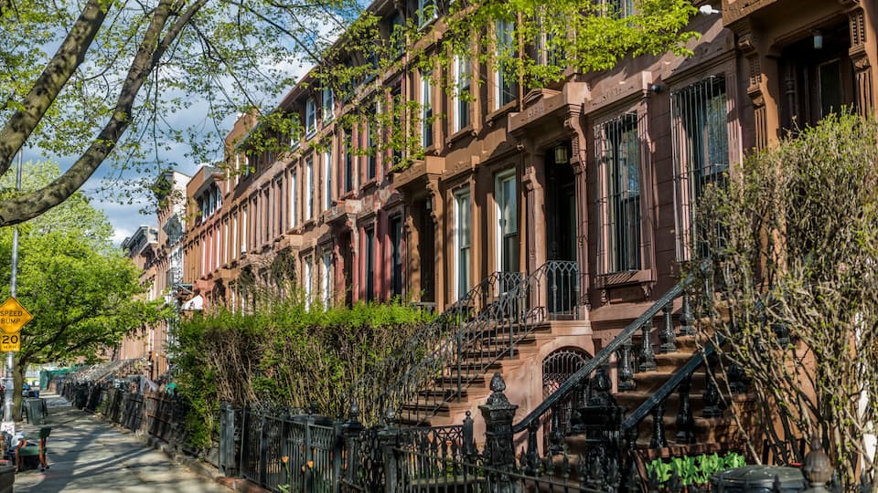 Fileira de casas em estilo brownstone com grades de ferro preto e placa indicando lombada na rua.