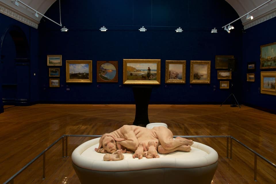 An art gallery with a central sculpture of a reclining figure surrounded by framed paintings on the walls.