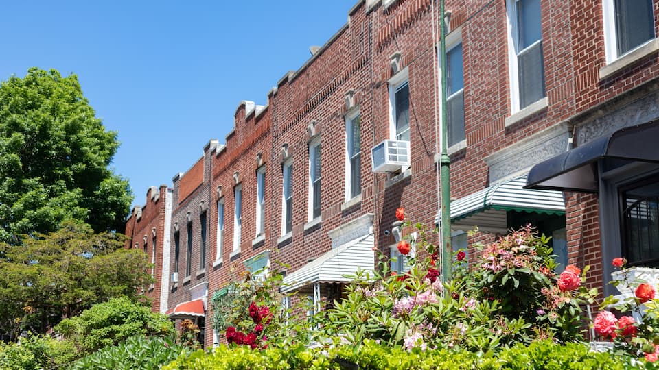 A row of red brick buildings with green awnings, surrounded by lush greenery and flowering plants.