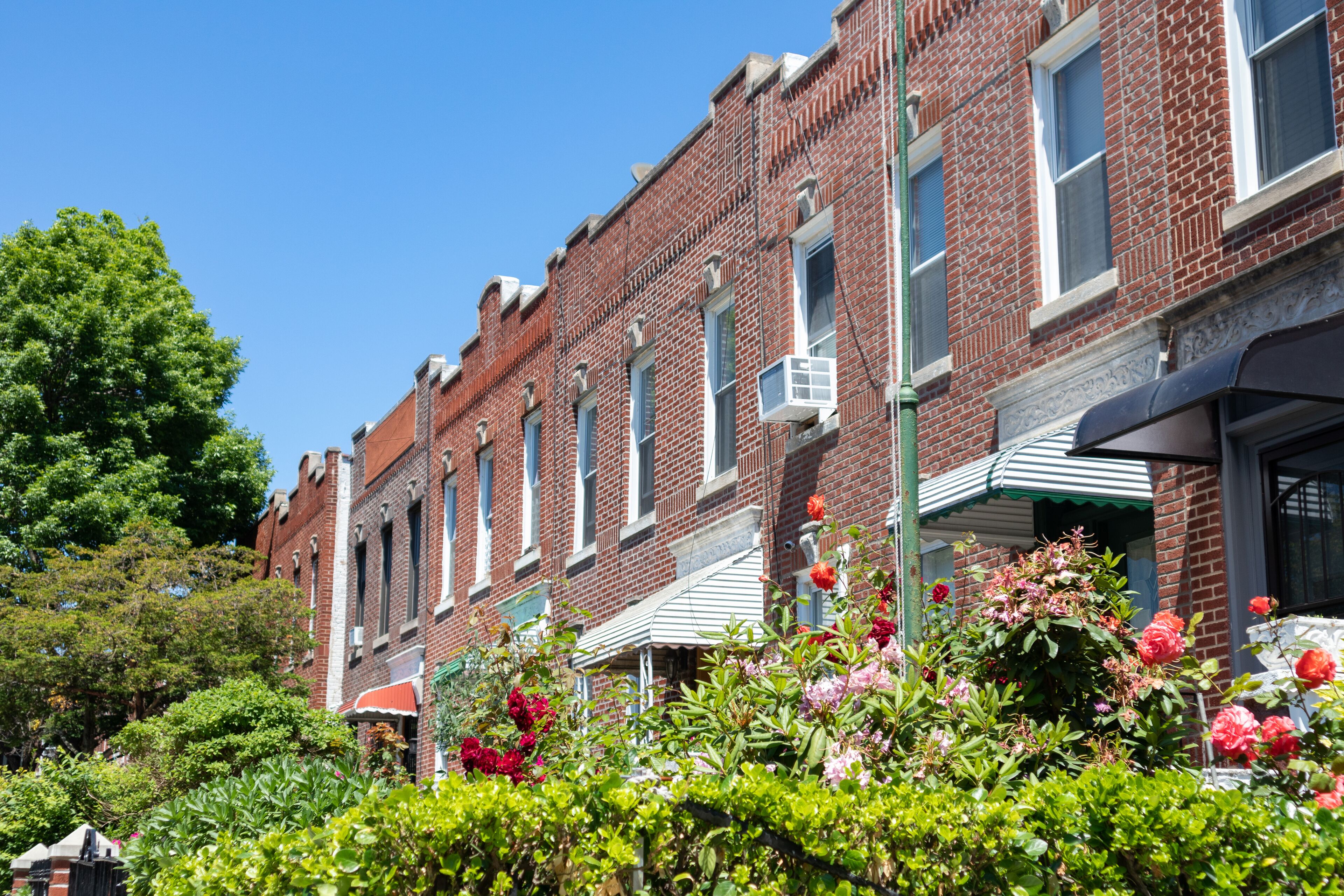 A row of red brick buildings with green awnings, surrounded by lush greenery and flowering plants.