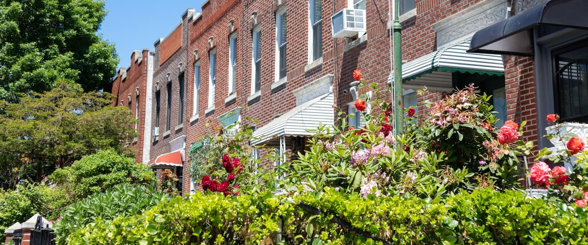 A row of red brick buildings with green awnings, surrounded by lush greenery and flowering plants.