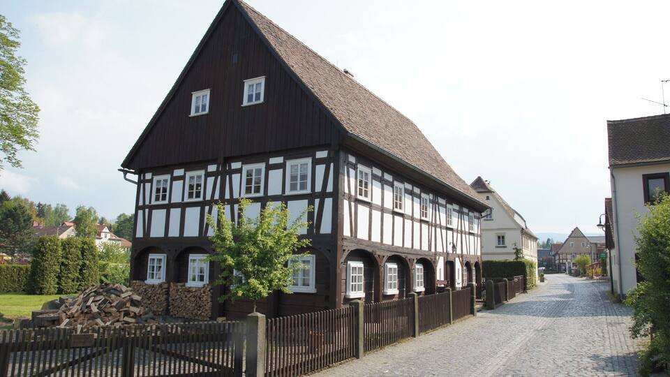 A traditional half-timbered house with a steep roof, a cobblestone street, and a wooden fence.