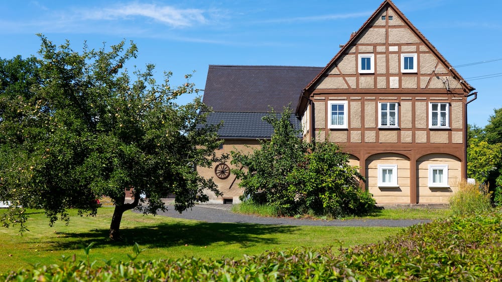 A traditional half-timbered house with a sloped roof, surrounded by lush greenery and trees.