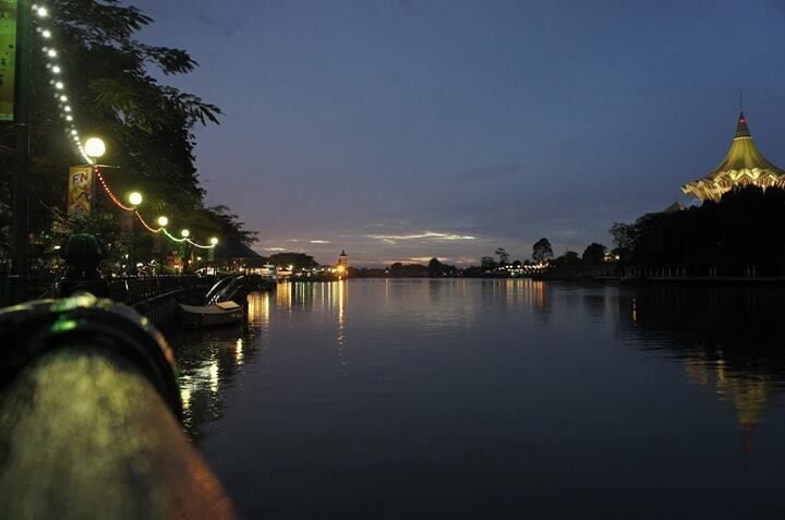 A calm river at dusk with a bridge, boats, and a lit-up building reflecting on the water.
