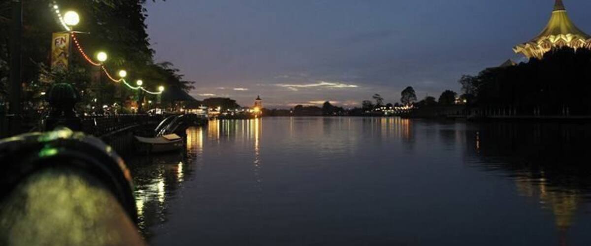 A calm river at dusk with a bridge, boats, and a lit-up building reflecting on the water.