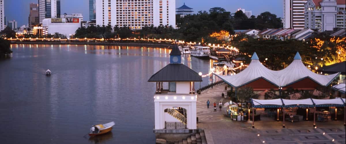 A waterfront cityscape with a prominent white building, a traditional pavilion, and a small boat on the water.