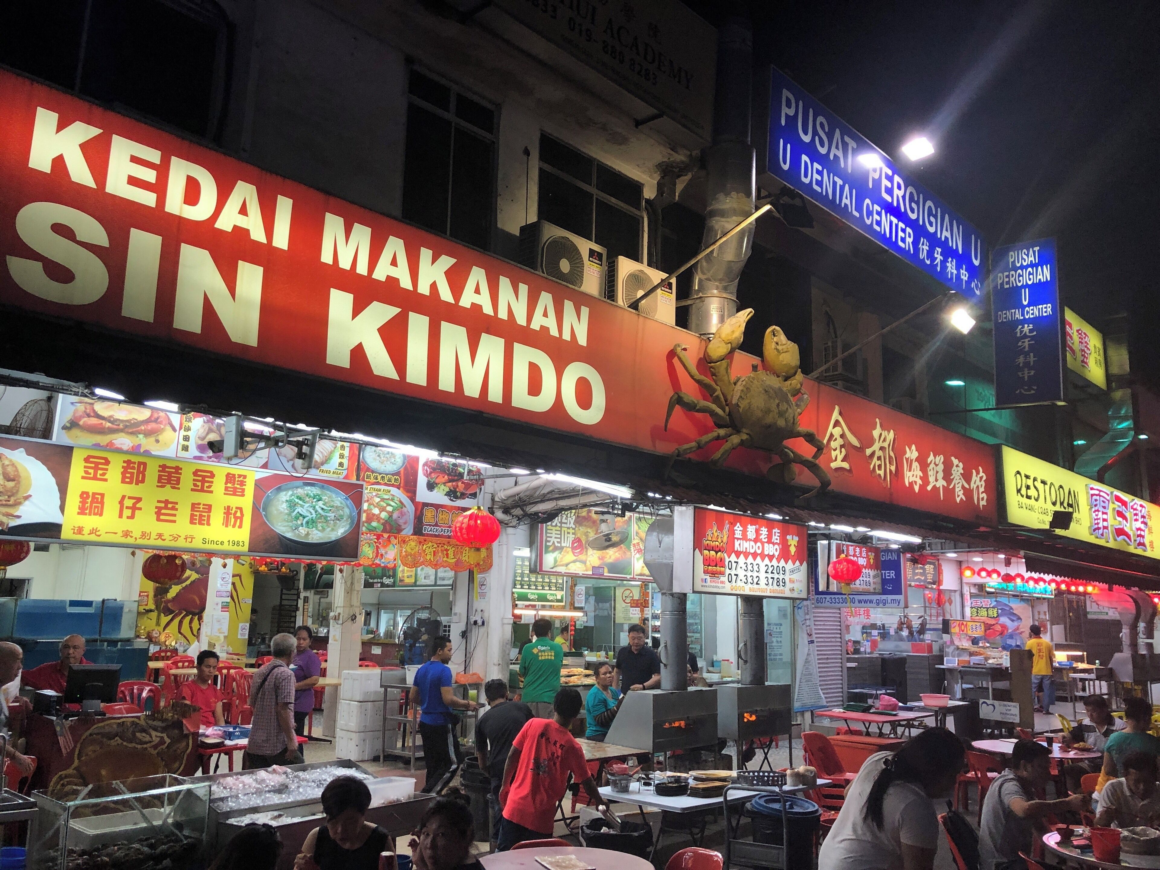 A bustling street food market with various food stalls, including a prominent sign for 'Kedai Makanan Sin Kimdo'.