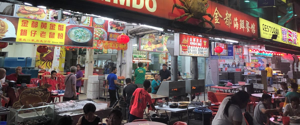 A bustling street food market with various food stalls, including a prominent sign for 'Kedai Makanan Sin Kimdo'.