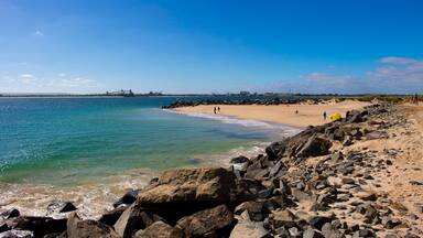 A rocky beach with clear blue water and a distant bridge.