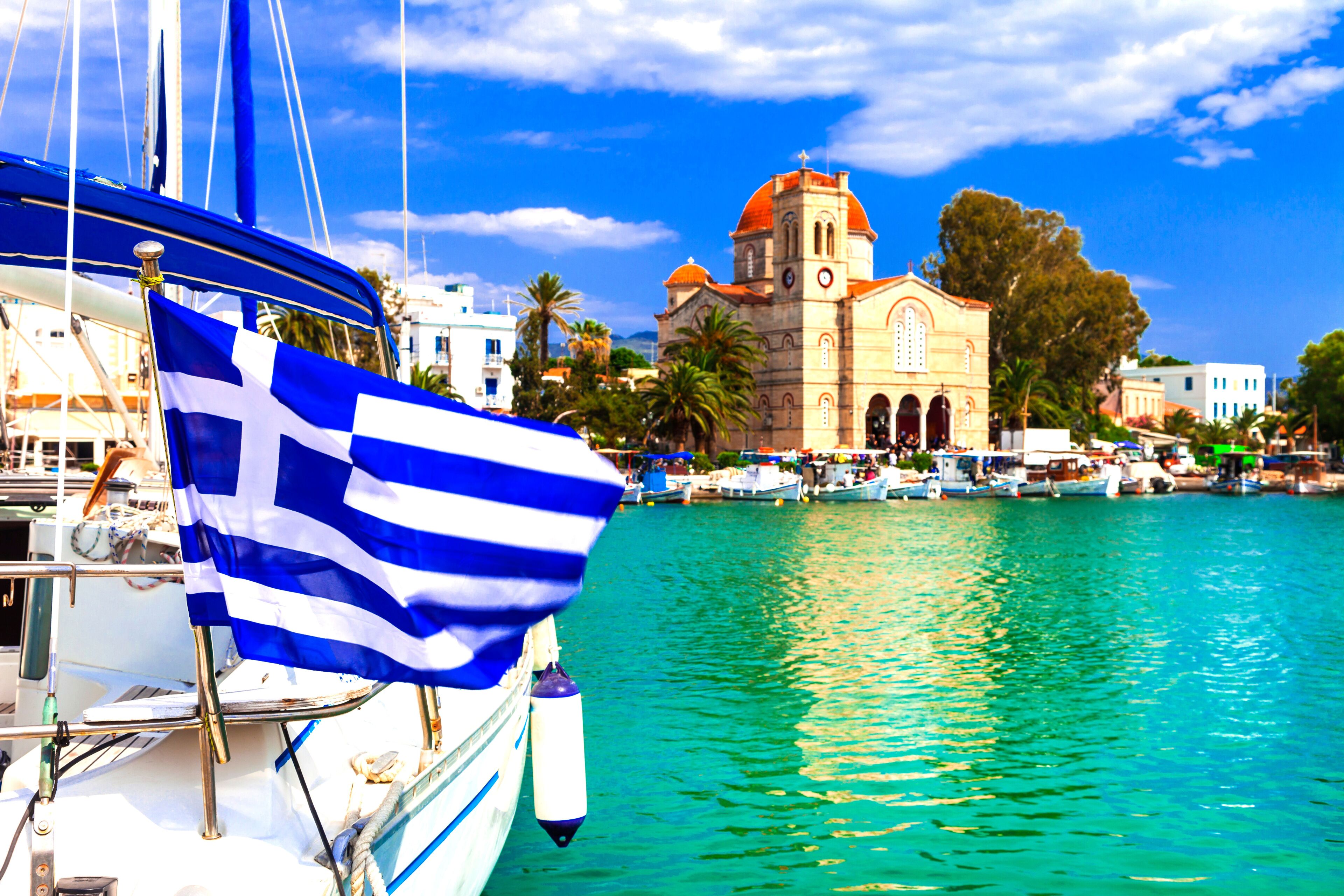 Cinematic View of Aegina Island Harbor — Greek National Flag Waving Over Luxury Yachts and Orthodox Church, Saronic Gulf Port Greece