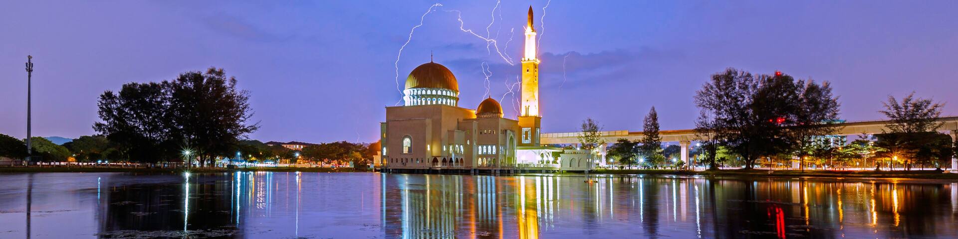 A mosque with a minaret and dome, reflecting on a calm body of water, with lightning in the sky.