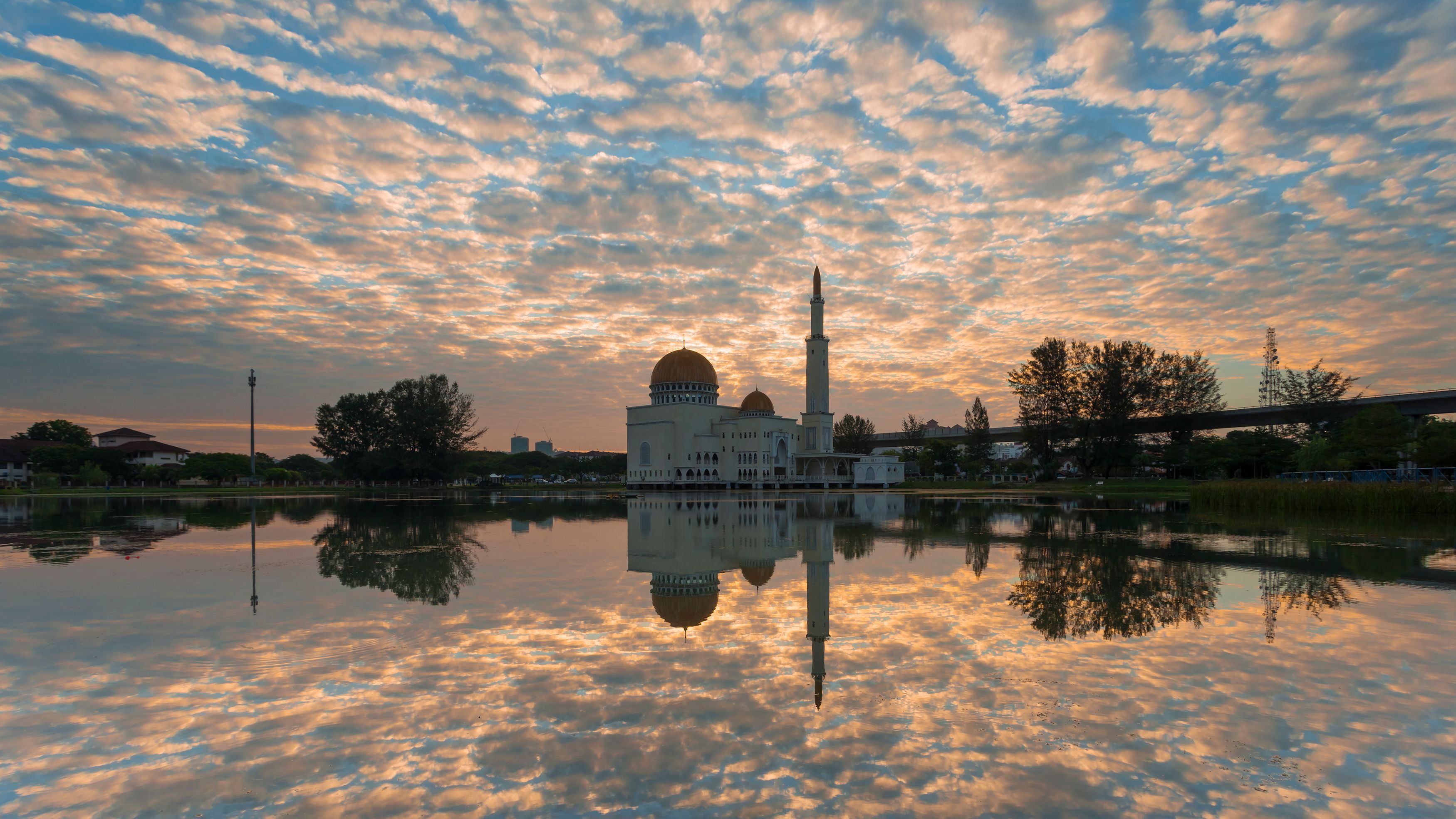 Una mezquita con un minarete alto y una cúpula, reflejada en una masa de agua tranquila bajo un cielo de atardecer.