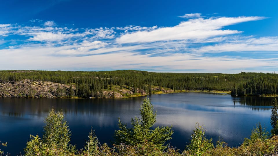 A calm lake surrounded by dense forest and rocky terrain under a clear blue sky.