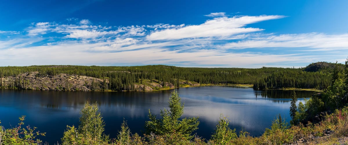 A calm lake surrounded by dense forest and rocky terrain under a clear blue sky.