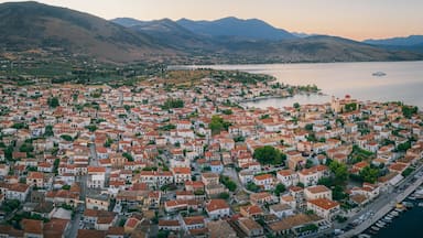 Uma vila costeira com edifícios de telhado vermelho, um porto de recreio e montanhas ao fundo.