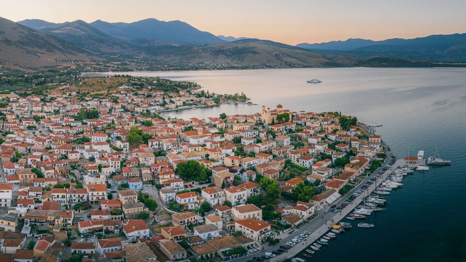 A coastal town with red-roofed buildings, a marina, and mountains in the background.