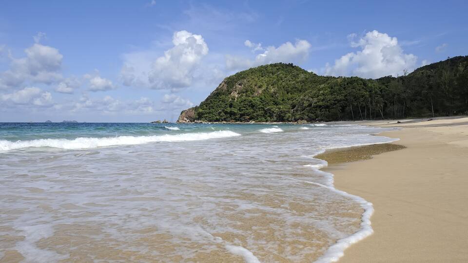 A sandy beach with gentle waves, a clear sky, and a forested hill in the background.