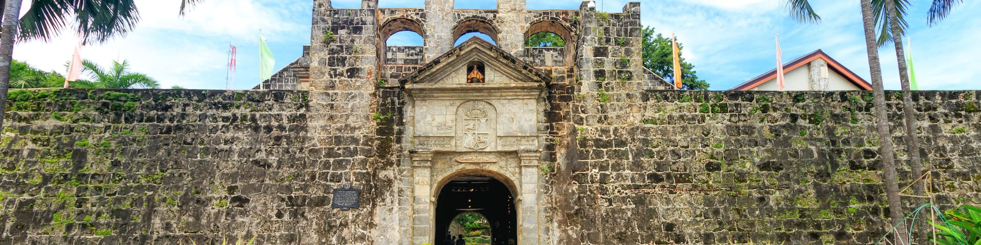 A historic stone gate with a central archway, flanked by two large potted plants, leading into a lush garden.