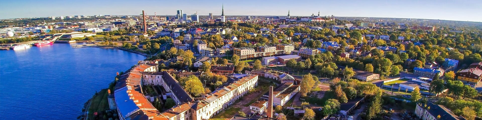 An aerial view of a coastal city with a prominent white building complex, a large body of water, and a distant city skyline.