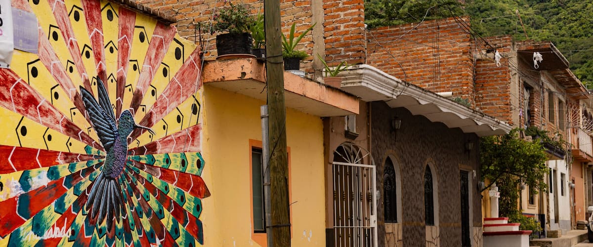 A colorful mural of a peacock on a building, a brick house with a balcony, and a cobblestone street.