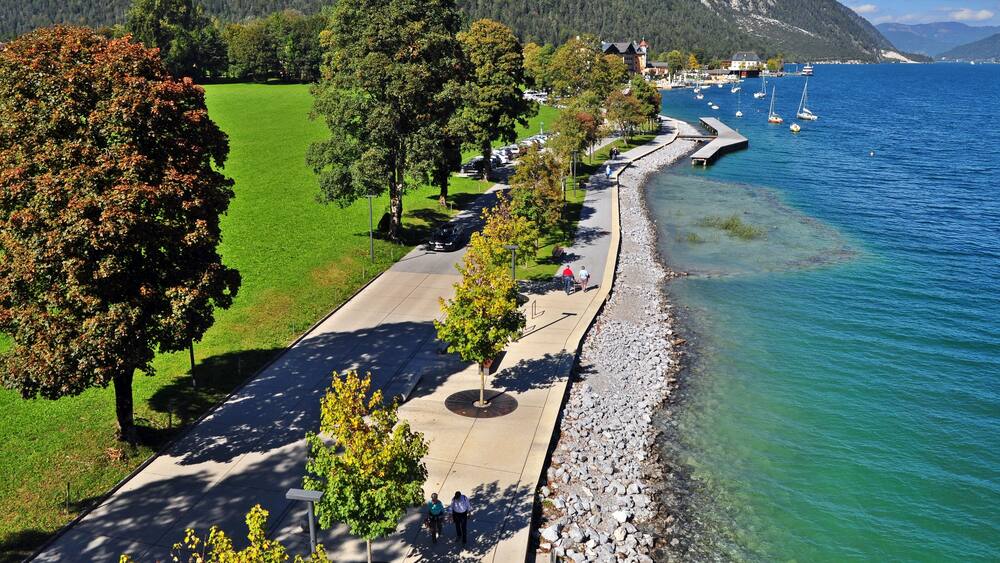 Eine Uferpromenade mit freiem Blick auf das Wasser, eine Baumreihe und ein ferner Gebirgszug.