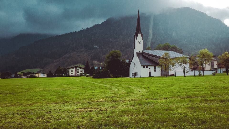 A church with a tall steeple in a grassy field with houses and mountains in the background.