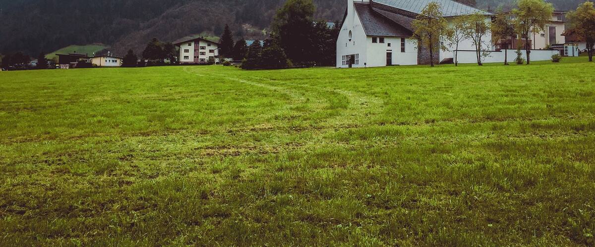 A church with a tall steeple in a grassy field with houses and mountains in the background.