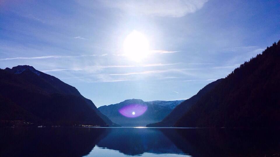 A calm lake with mountains on either side and a bright sun in the center.