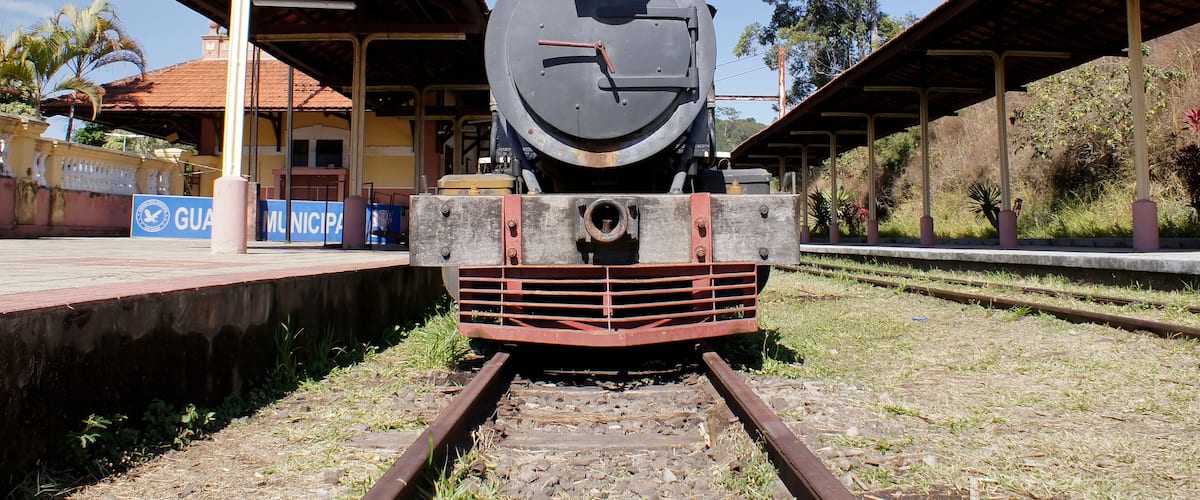 A vintage steam locomotive at a railway station with a covered platform and a sign reading "GUAM MUNICIPAL".