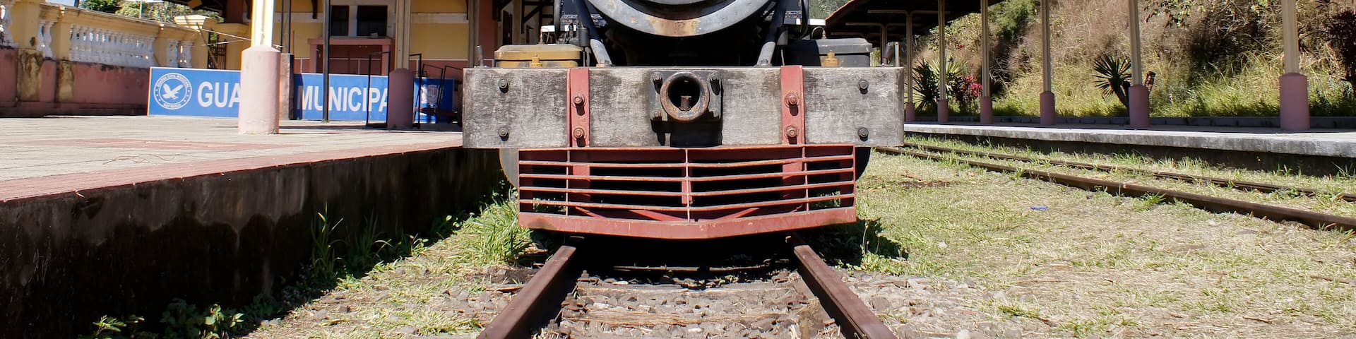 A vintage steam locomotive at a railway station with a covered platform and a sign reading "GUAM MUNICIPAL".