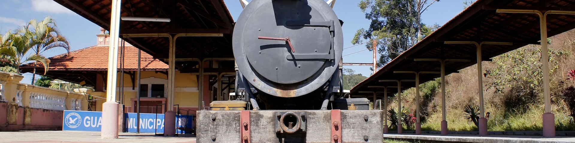 Una locomotora de vapor antigua en una estación de tren con una plataforma cubierta y un cartel que dice "GUAM MUNICIPAL".