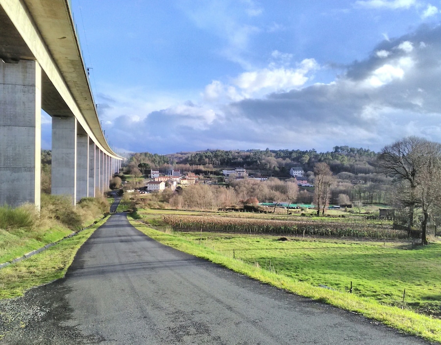 A rural road under a bridge, surrounded by fields and houses.