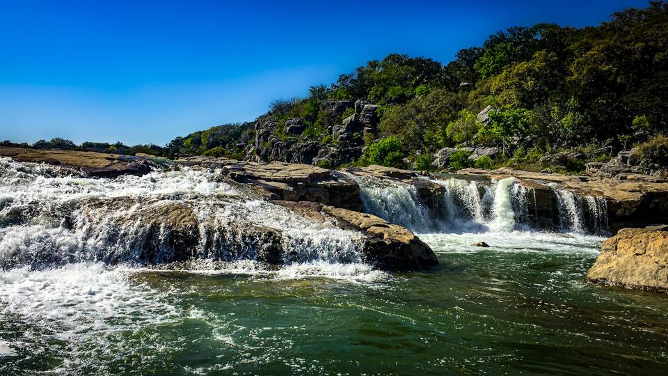 A multi-tiered waterfall cascading over rocky terrain with dense forest in the background.