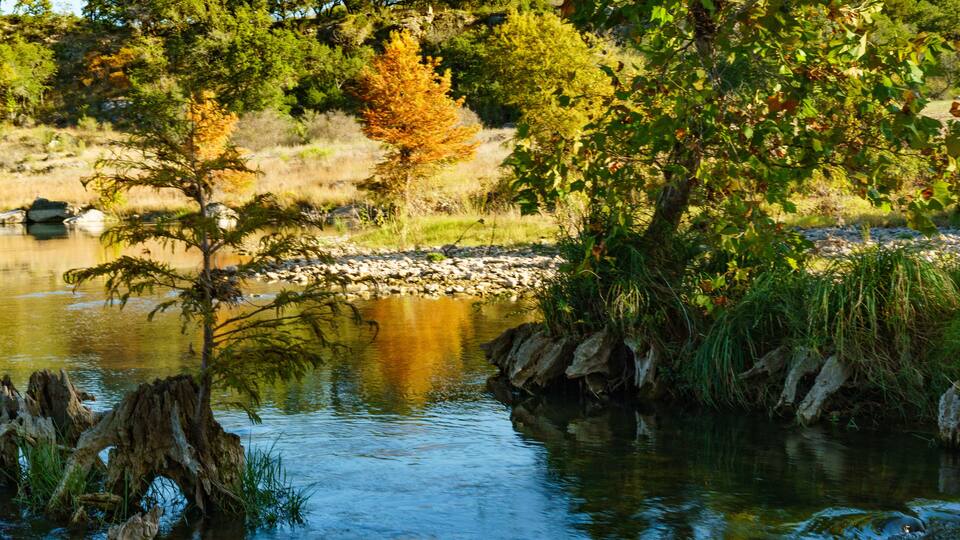 A calm river with trees reflecting on the water, surrounded by rocks and greenery.