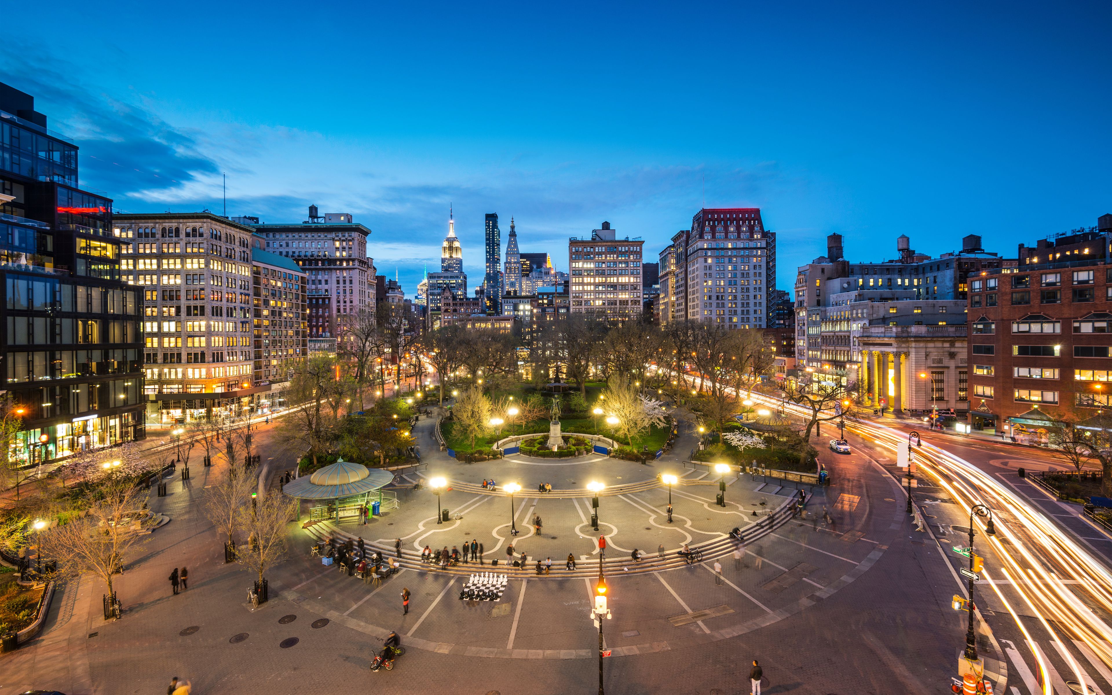 A cityscape at dusk with illuminated buildings, a central park with a fountain, and a busy intersection with light trails from moving vehicles.