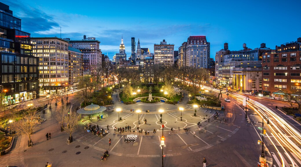 A cityscape at dusk with illuminated buildings, a central park with a fountain, and a busy intersection with light trails from moving vehicles.