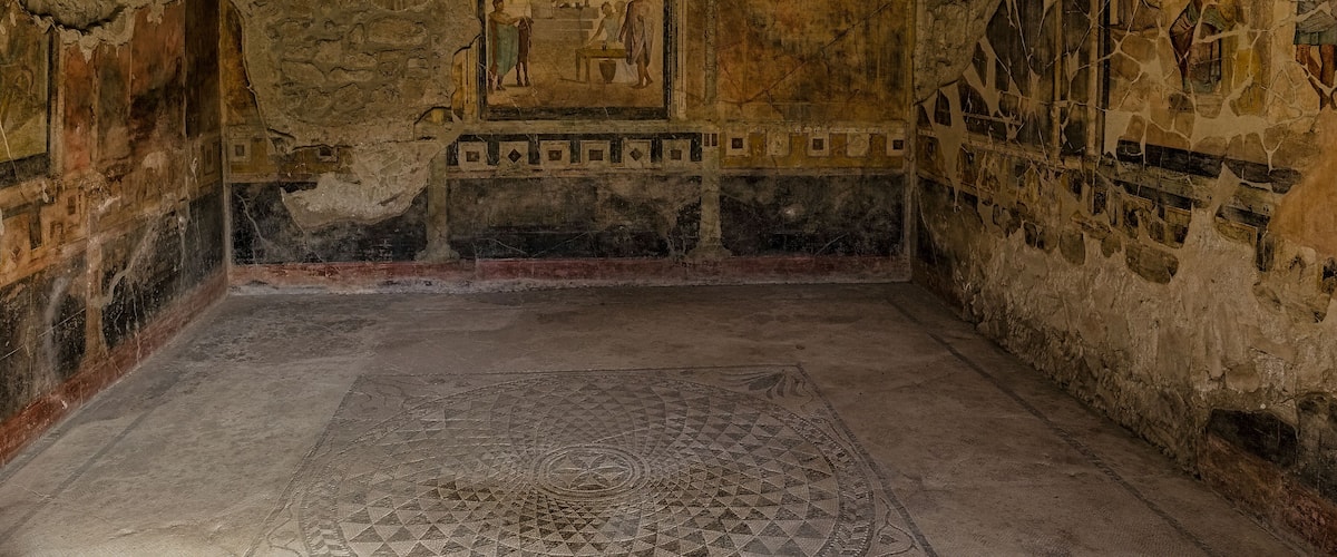 An ancient room with frescoes, a geometric floor pattern, and stone walls.
