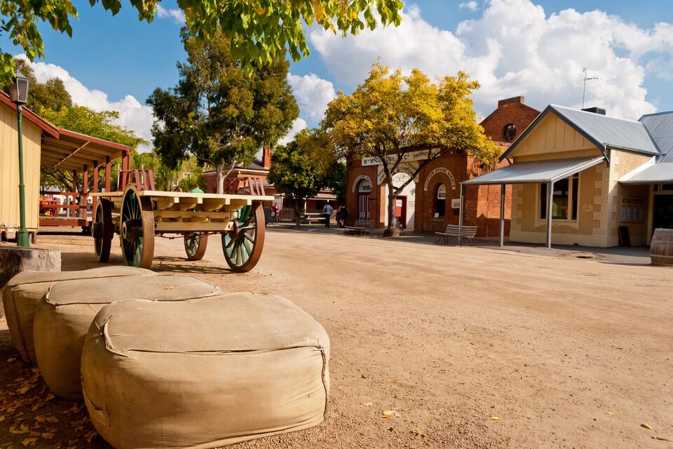 Ein historischer Stadtplatz mit einem hölzernen Wagen, großen Säcken und historischen Gebäuden.