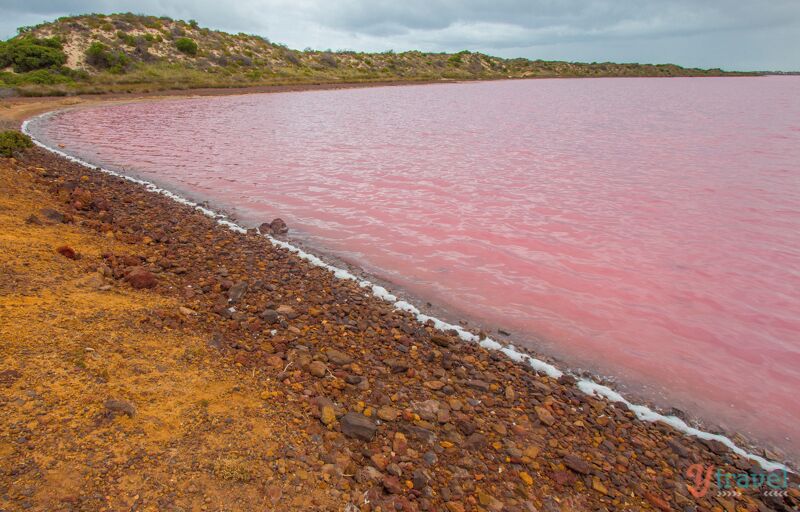 A beach with pink water, pebbles, and a hilly landscape.
